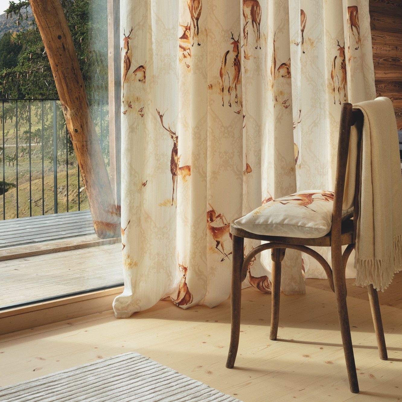 Manufaktur für individuelle Raumgestaltung Holzstuhl vor einem Vorhang mit Hirschmotiv an einer Glastür mit Bergblick